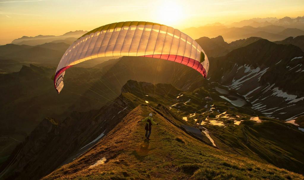 Paraglider launching from a mountain ridge at sunset, with the wing overhead and alpine terrain below.